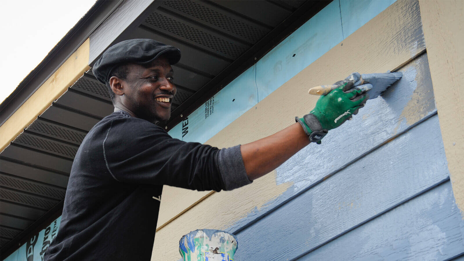 A man smiling as he brushes light blue paint on a tan house, just below the roof. He wears a black long-sleeved shirt, a black hat, and green work gloves. There is a can of paint below his raised arm.