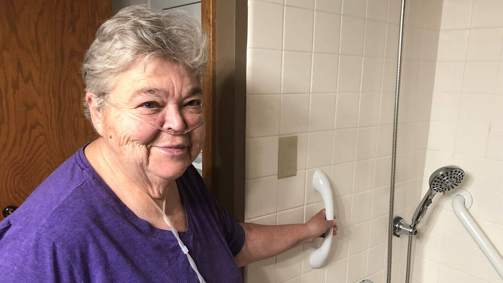 Linda smiling in a purple shirt in her bathroom, showing off her new shower grab bars and handheld shower head.
