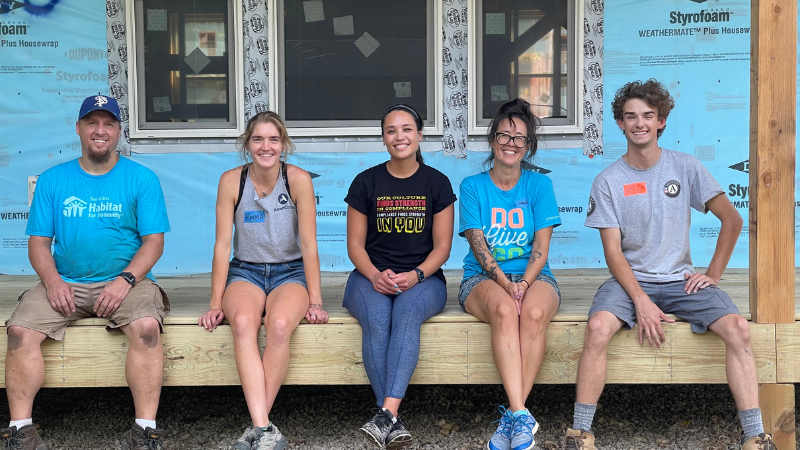 From left to right, sitting on the porch of a home under construction: Regular Volunteer Corey, Emma, two volunteers, and AmeriCorps member Nick.