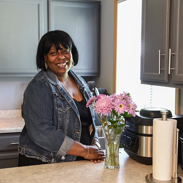 Angela smiling in her kitchen next to a counter with a bouquet of pink flowers.