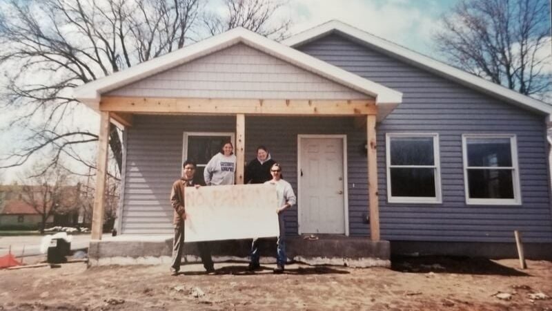 Standing outside the front porch of an in-progress gray house, two site supervisors hold a handmade no parking sign. On the right is Heather's site supervisor Pete Brownlee . Behind him is fellow AmeriCorps Carrie, and to the left of her is Heather. All are in sweatshirts and there are no leaves on the trees.