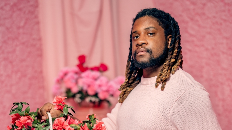 Denzel Belin, a Black man, posing in front of a pink background and holding a basket of pink flowers.