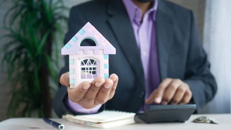 Close up of man in a suit holding a miniature house in his outstretched hand.