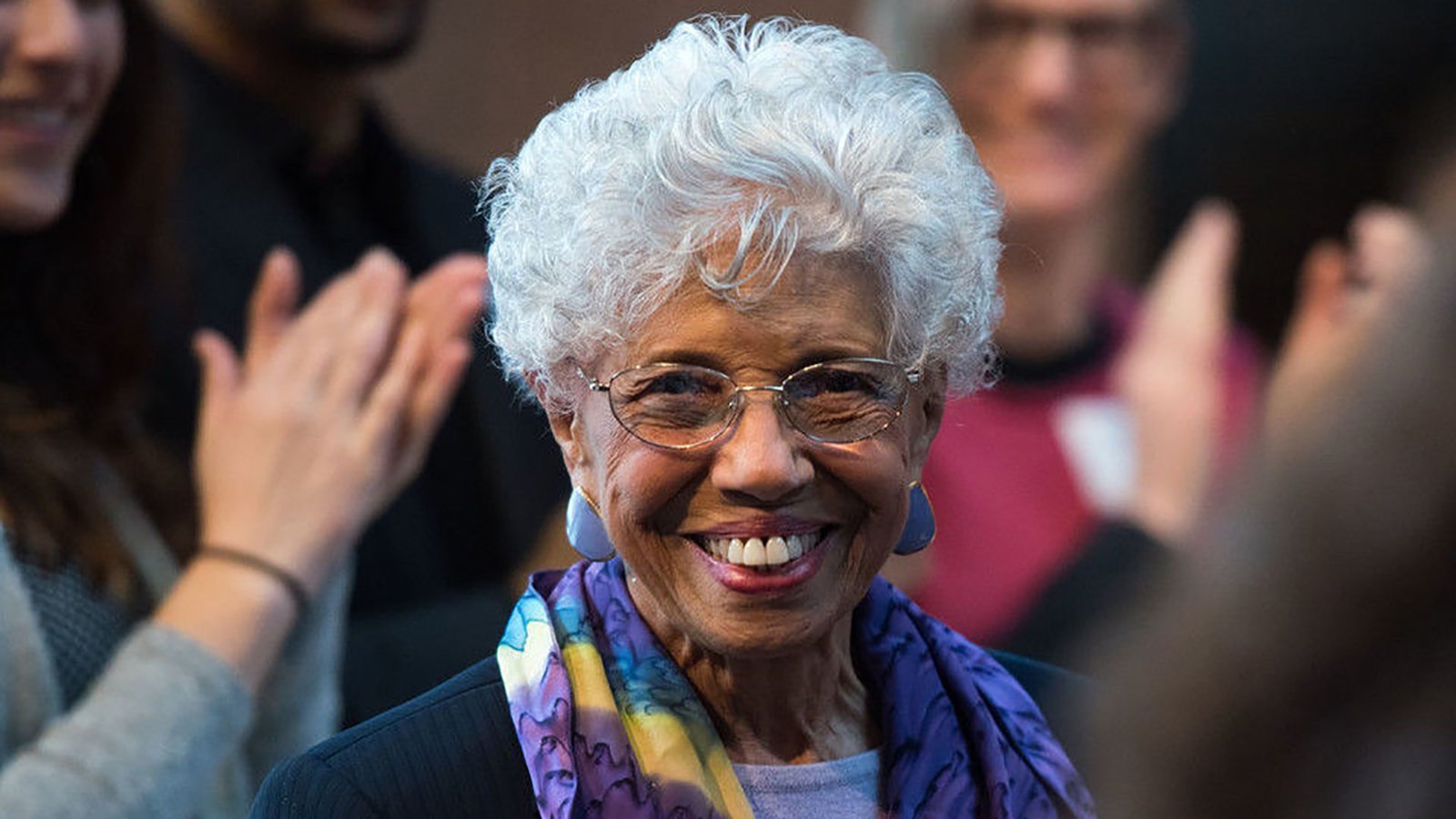 A close-up on Dr. Josie Johnson, in a purple shirt, black pinstripe jacket, and a blue, purple and yellow patterned infinity scarf, smiling in a crowd with her hands clasped. 