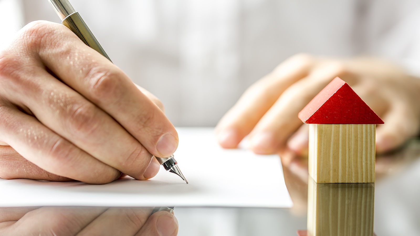 Brightly lit, and on a reflective table, one hand writing on a sheet of white paper with a fountain pen, with the other hand resting on the edge of the paper. A small square wooden block sits next to the paper, with a red triangle on top to form the shape of a house.