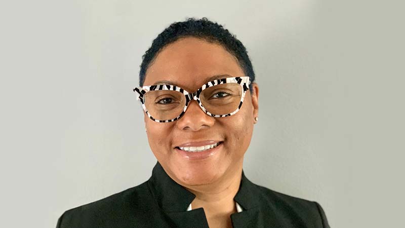 Shereese Shereese Turner smiling in front of a gray background, wearing a white shirt, black formal jacket, and black and white striped glasses.