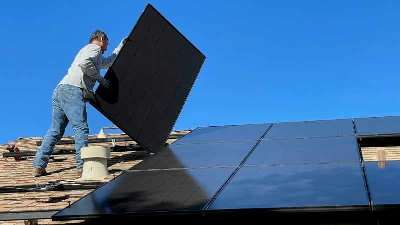 Man on a roof installing a large solar panel against a blue sky.