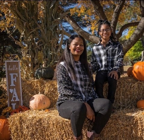 A Black mother and daughter pose at a Halloween display with straw bales.