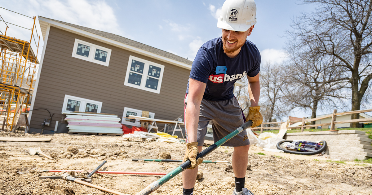Jesse Mamaril in a US Bank shirt and construction hat and gloves, digging dirt on a build site.