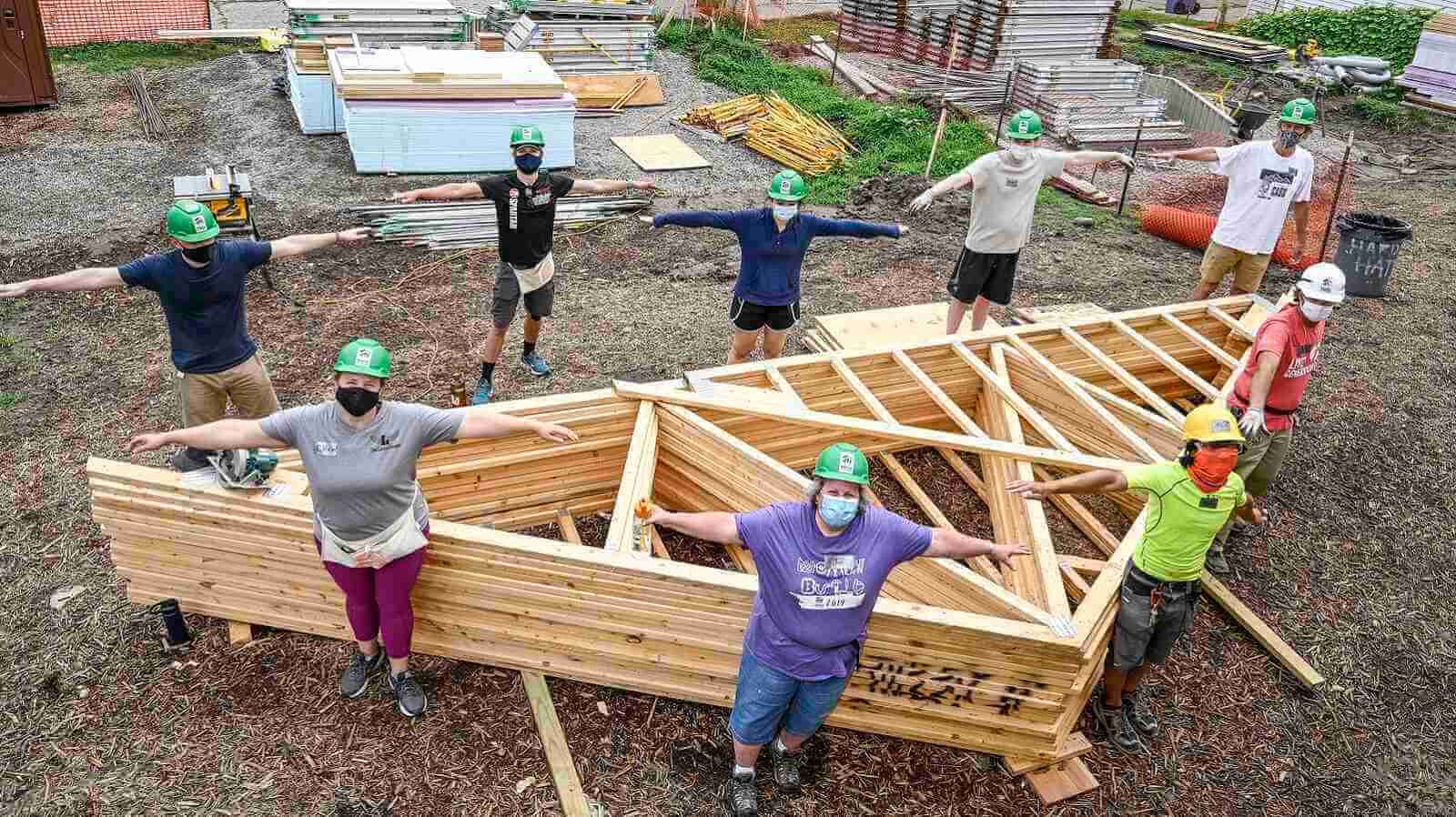 Nine volunteers demonstrating social distancing with open arms as they stand around a series of wood beams forming the triangular roof of a house.
